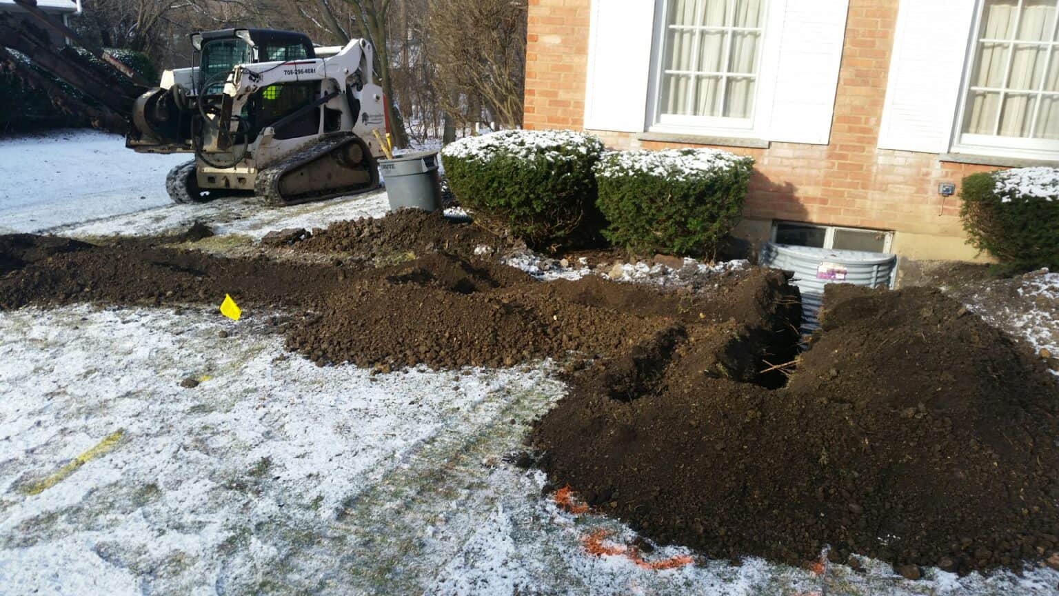 Skid steer digging along a brick home’s foundation for new window wells